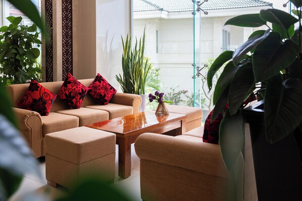 Una sala de estar con un sofá color beige, un reposapiés a juego y una mesa de centro de madera. Hay varias plantas en macetas y un jarrón con flores sobre la mesa.