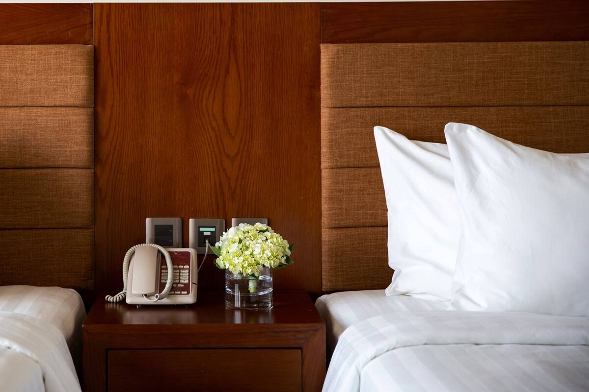 Une chambre d’hôtel avec deux lits, des têtes de lit en bois, une table de chevet avec un téléphone, un vase avec des fleurs et une horloge numérique.