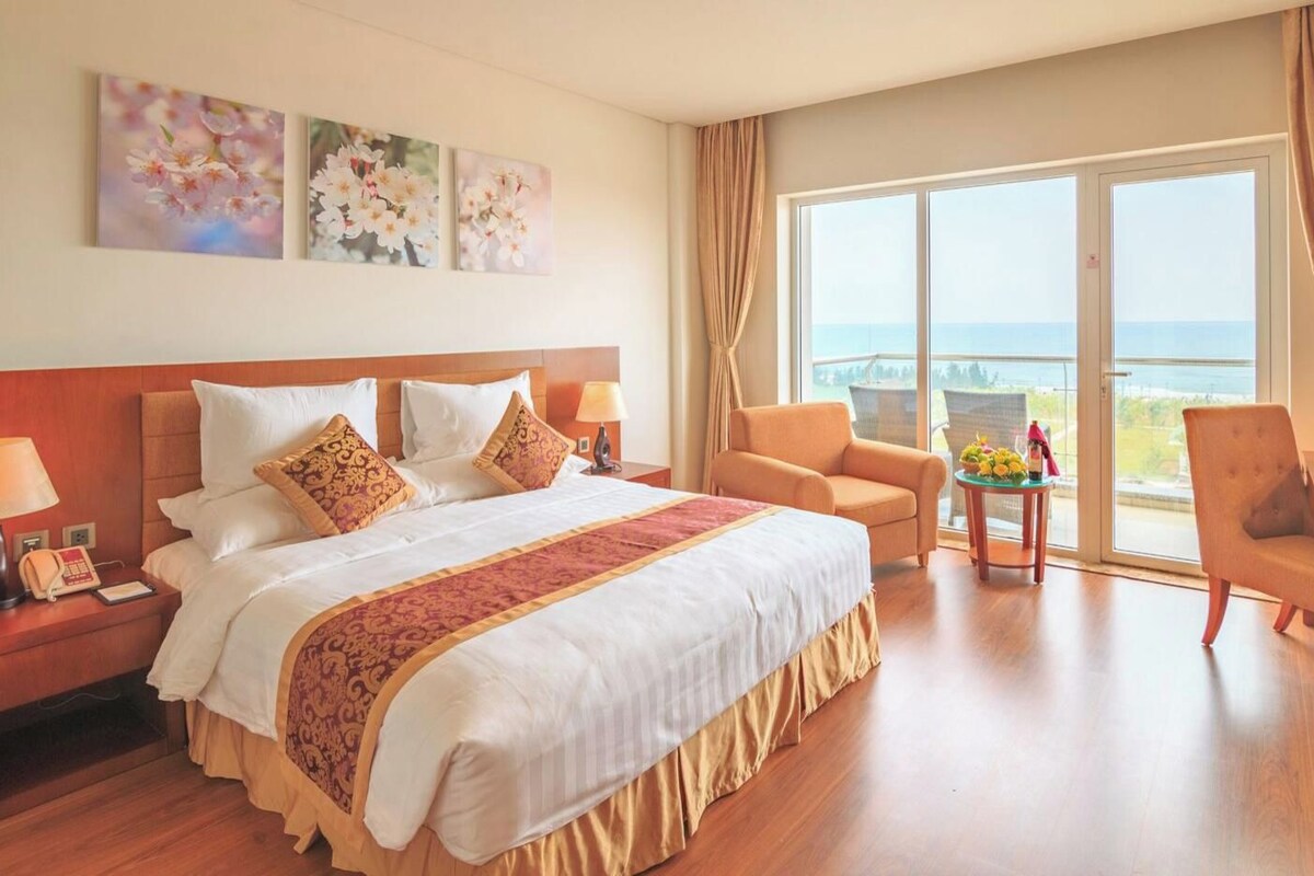 A hotel room with a large bed, a desk, a chair, and a view of the sea.