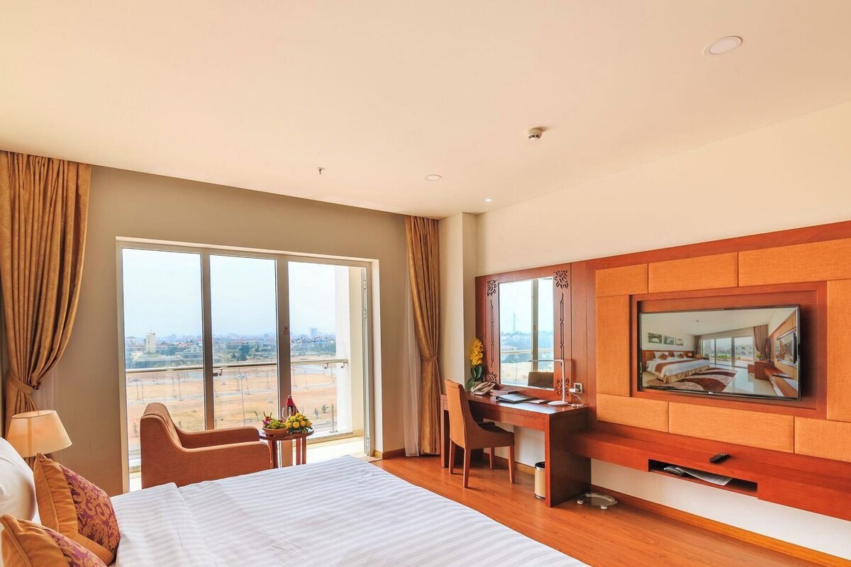 A hotel room with a large bed, a desk with a chair, a television, and a view of the city.