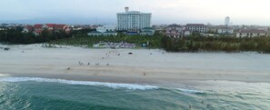 A beach resort with a large building in the background, a sandy beach, and ocean waves. - Gold Coast Hotel Resort & Spa (Dong Hoi)