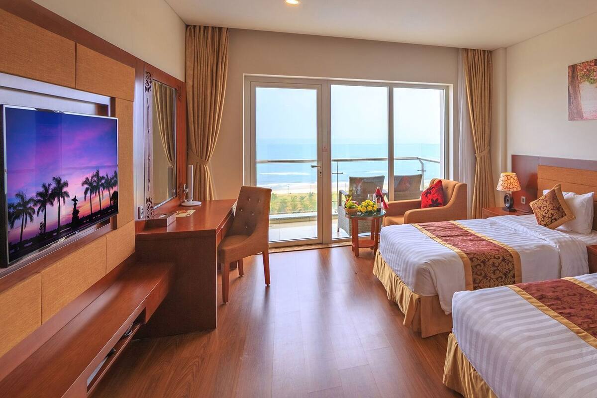 Une chambre d’hôtel équipée d’une télévision, de deux lits, d’un bureau, d’une chaise, d’un balcon avec vue sur la mer et d’œuvres d’art accrochées au mur.
