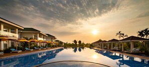 Una piscina con sillones de descanso y sombrillas, rodeada de edificios con vista despejada al atardecer.