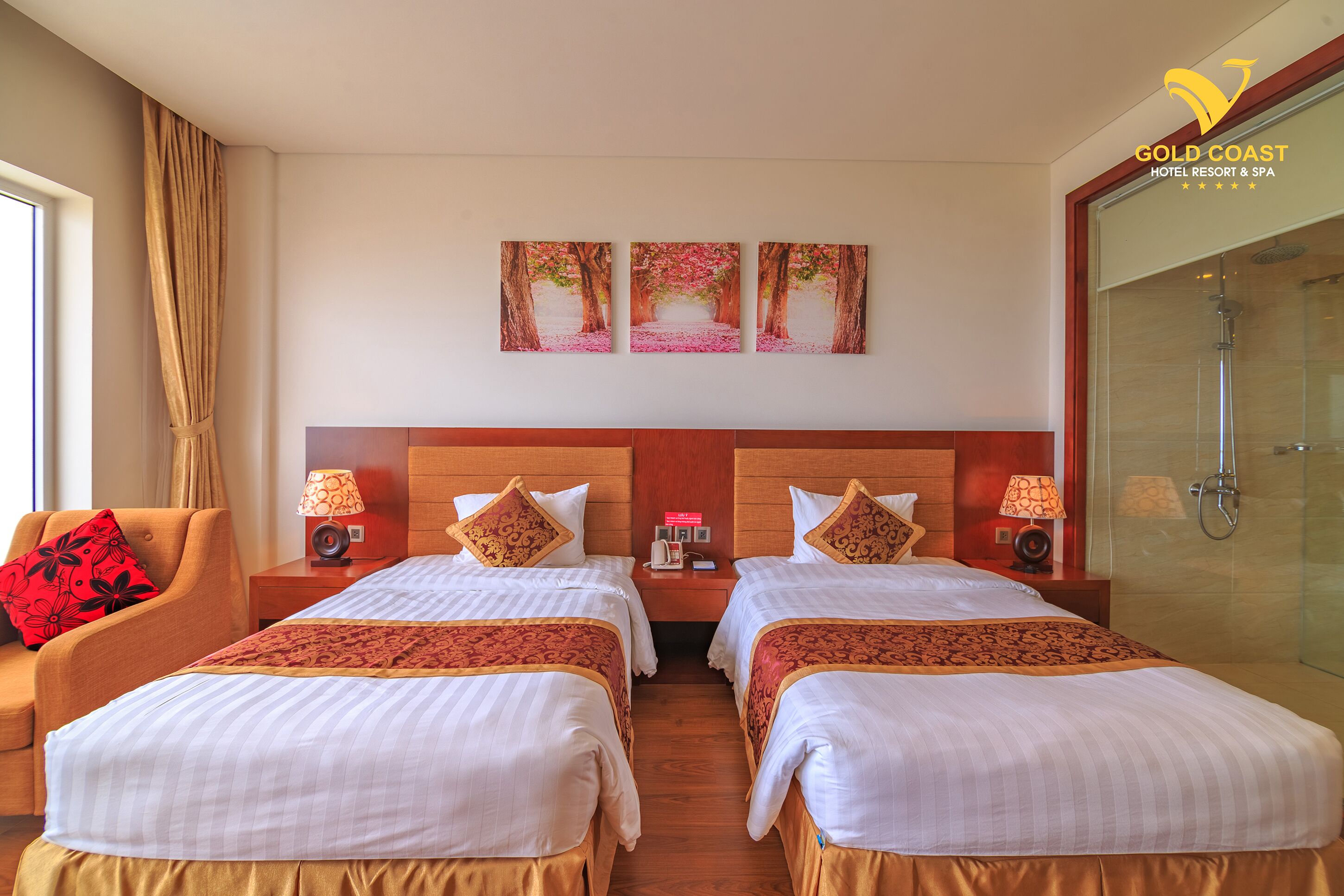 A hotel room with two beds, a wooden headboard, a glass shower, and a red and white patterned wall hanging.