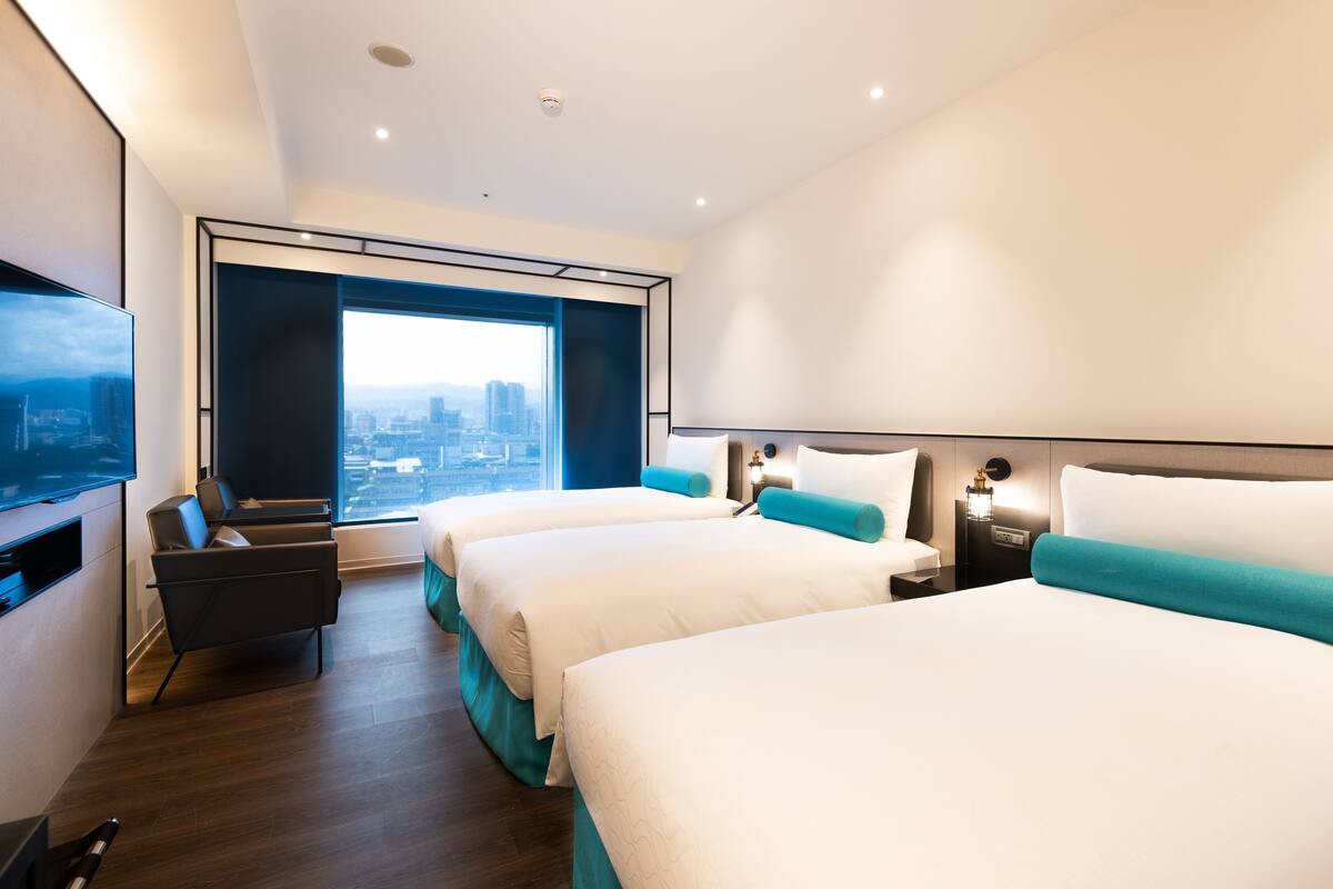 A modern hotel room with two beds, a TV, a desk, and a large window offering a city view.