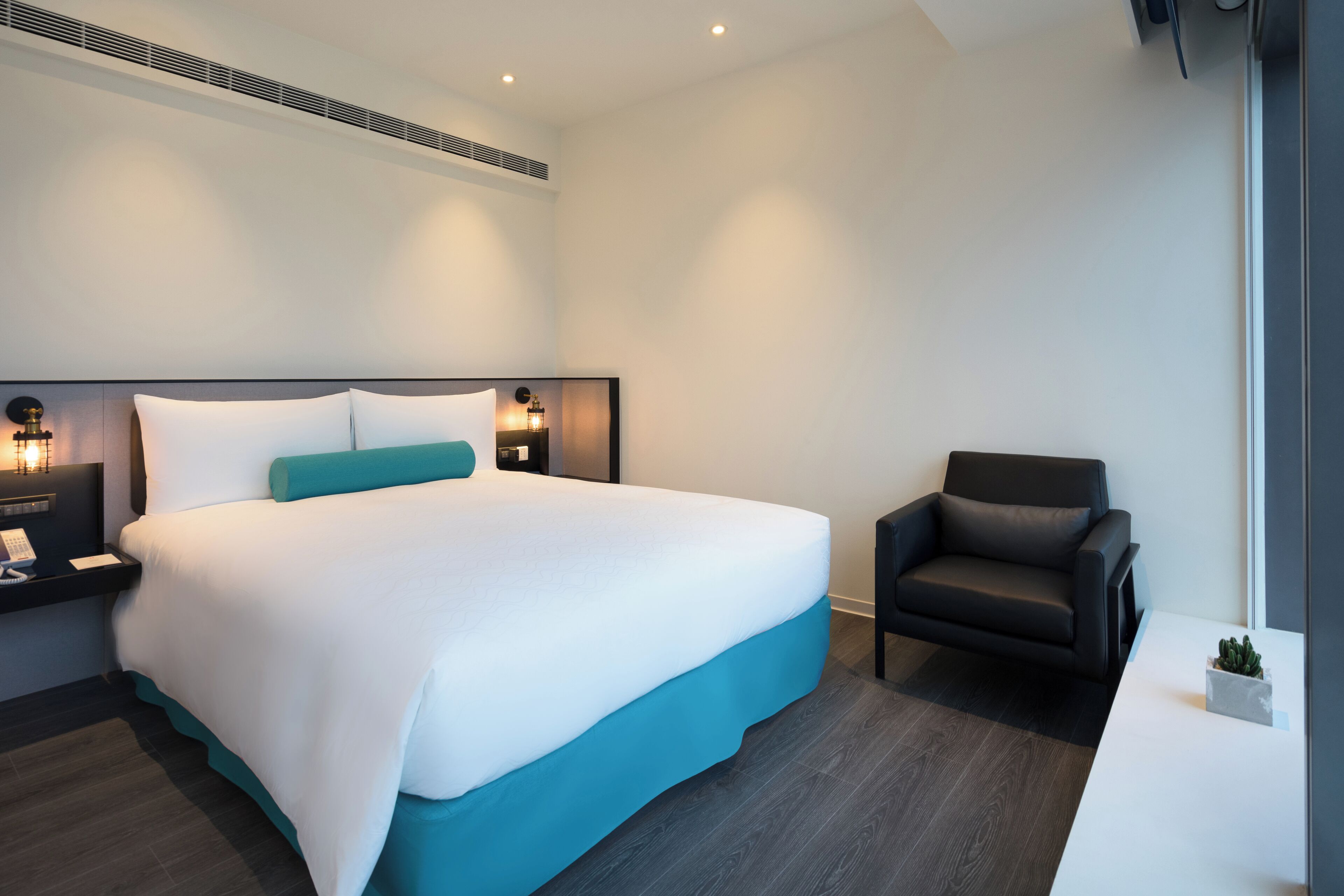 Quarto de hotel moderno com uma cama grande, uma cadeira, uma mesinha de cabeceira e uma pequena planta.