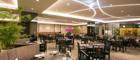 A spacious dining area with multiple tables and chairs, a central floral arrangement, and a modern ceiling design.