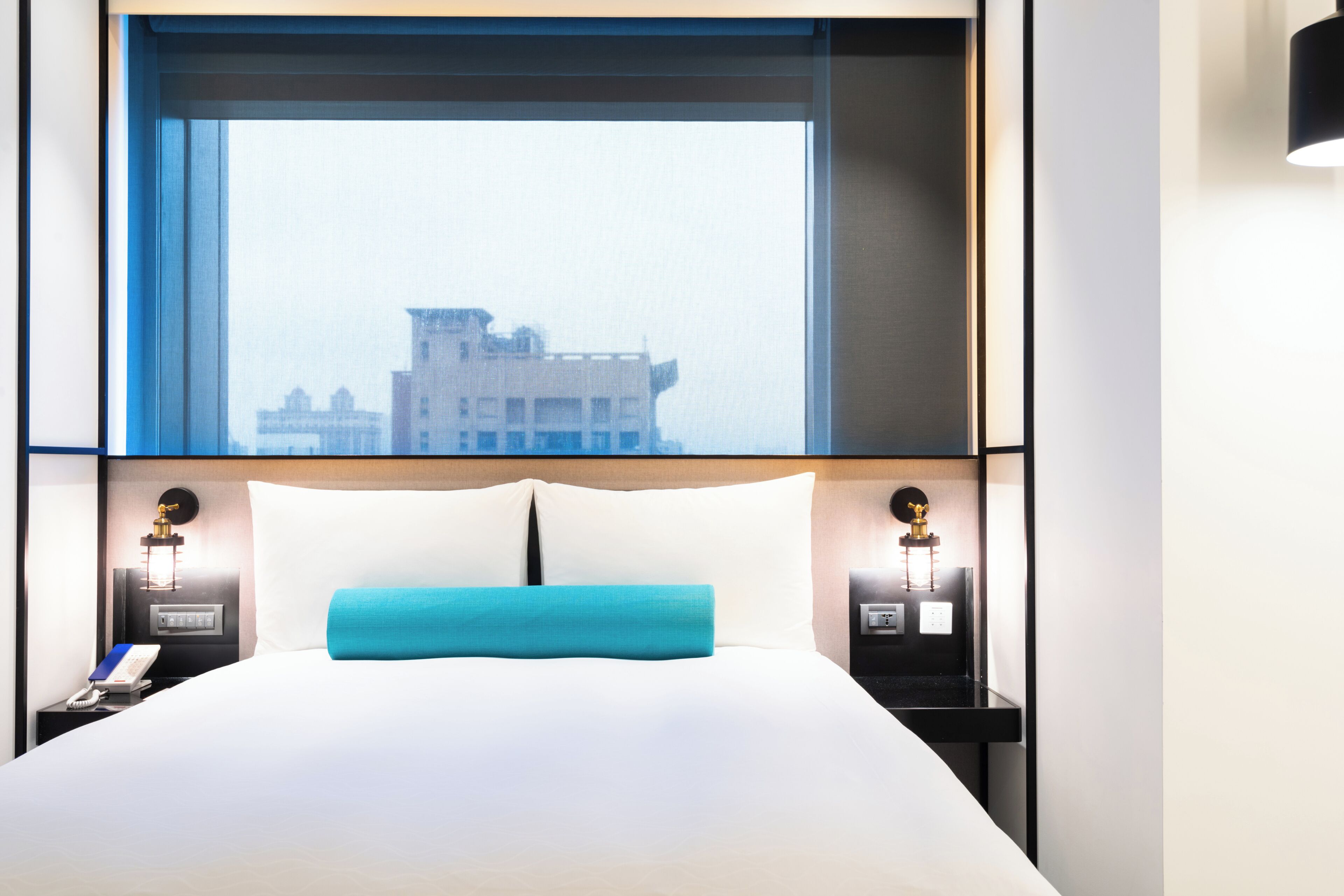 A hotel room with a large bed, two bedside tables with lamps, a window with a view of a building, and a nightstand with a phone and a book.