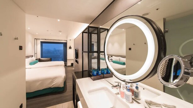 Une chambre d’hôtel moderne avec un grand lit, une salle de bain dotée d’un grand miroir rond et une vue sur la ville par une grande fenêtre.