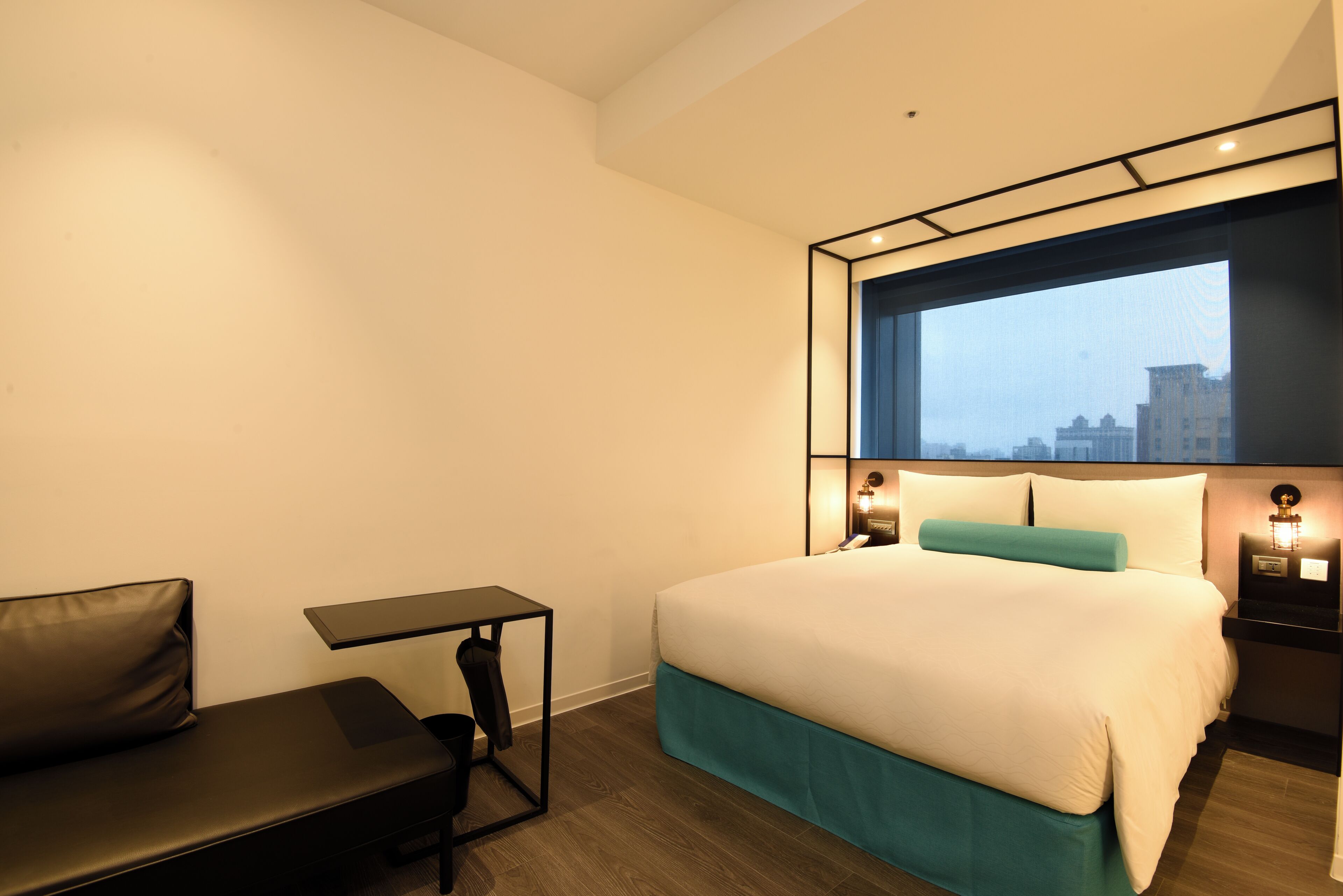A hotel room with a bed, a small table, a bench, and a window with a city view.