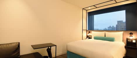 A hotel room with a bed, a small table, a bench, and a window with a city view.