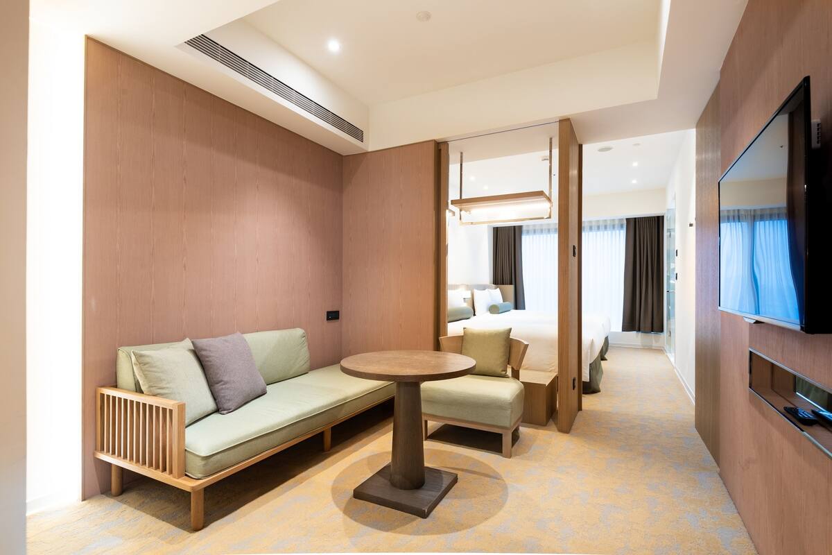 A modern hotel room with a sofa, a small round table, a bed, a TV, and a mirror.