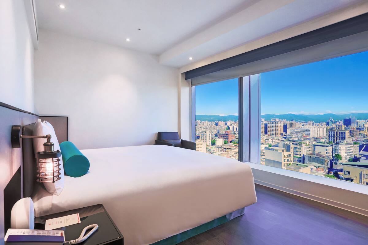 A modern hotel room with a large bed, a nightstand, a lamp, and a view of a cityscape through large windows.