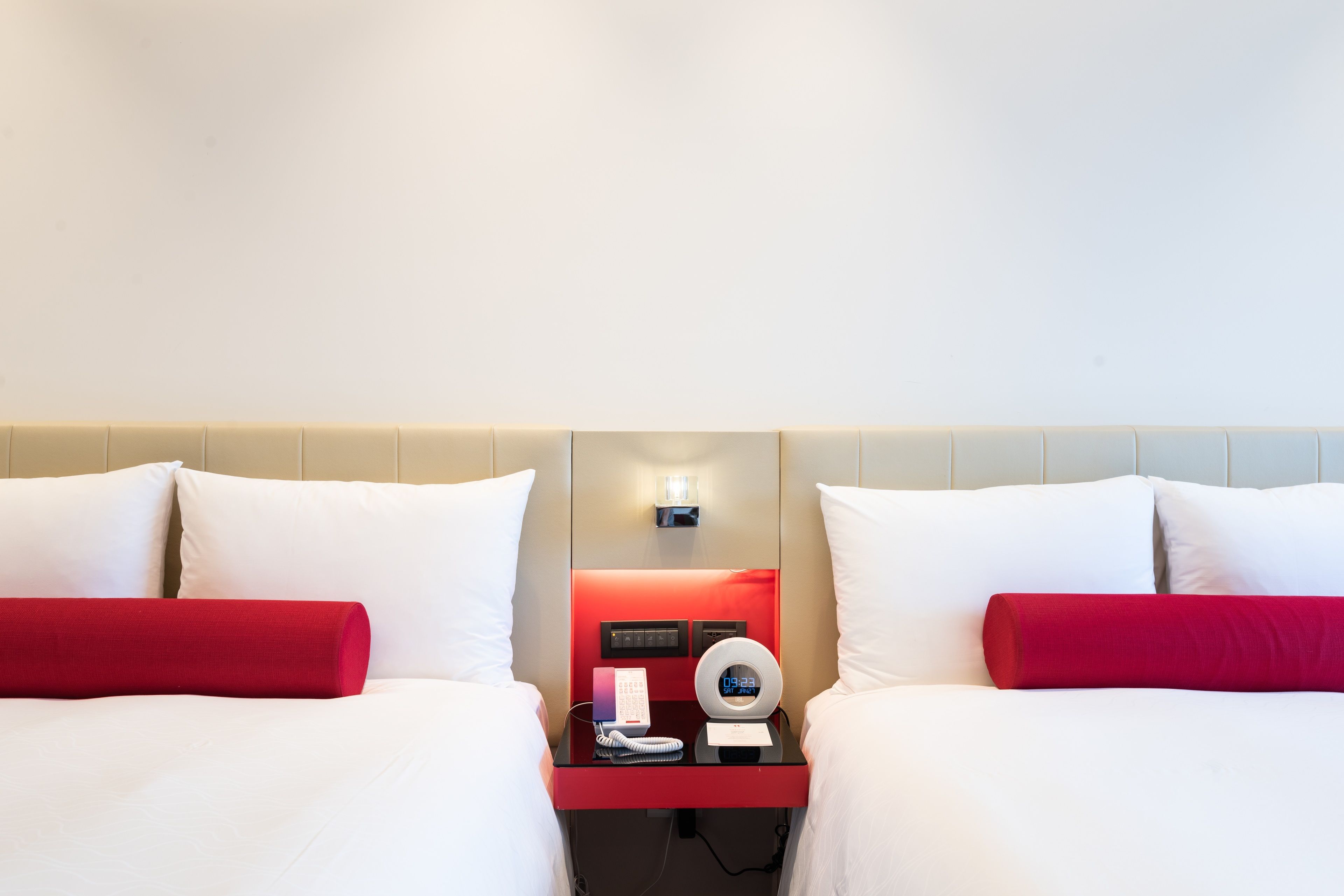 Habitación de hotel con dos camas, cada una con ropa blanca y una almohada roja decorativa. Hay una mesita de noche entre las camas con un teléfono, un reloj digital y una lámpara pequeña.