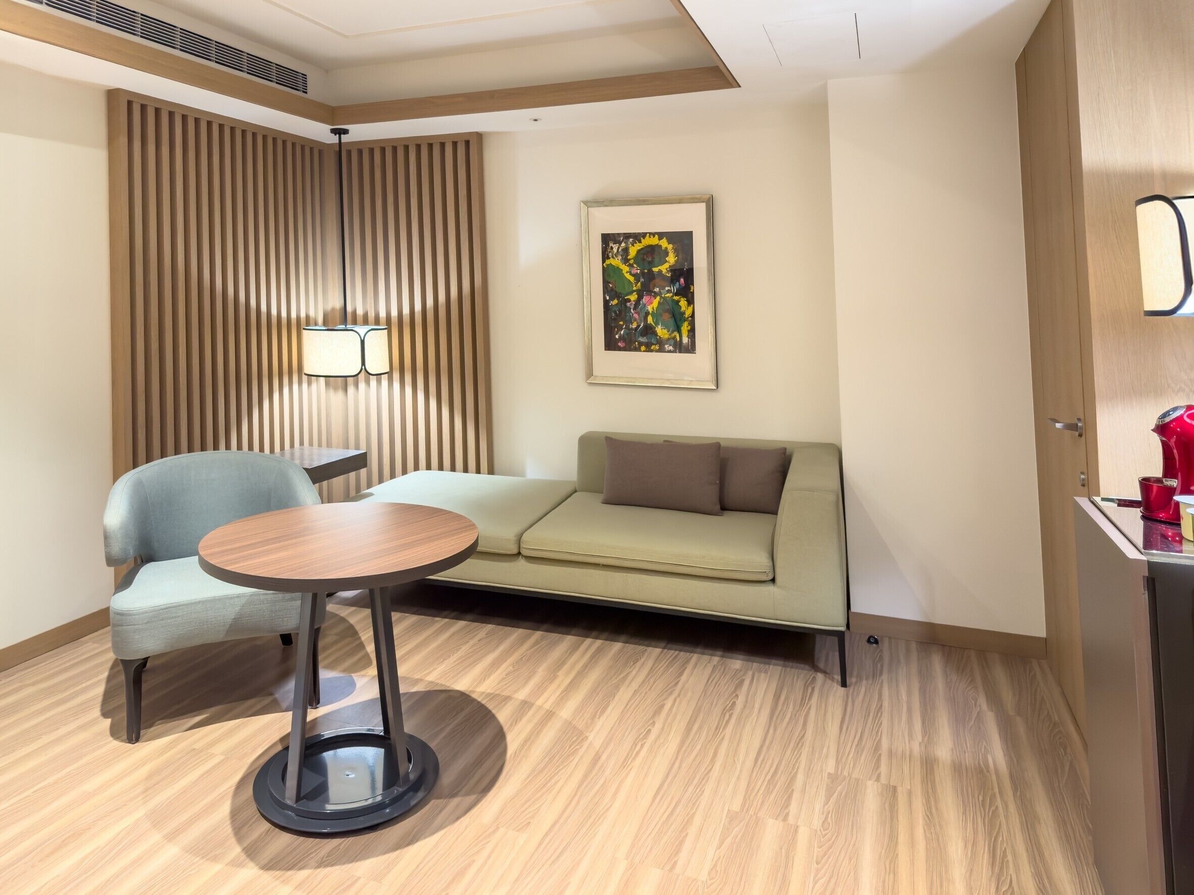 A hotel room with a wooden floor, a sofa, a chair, a small table, a lamp, and a painting on the wall.