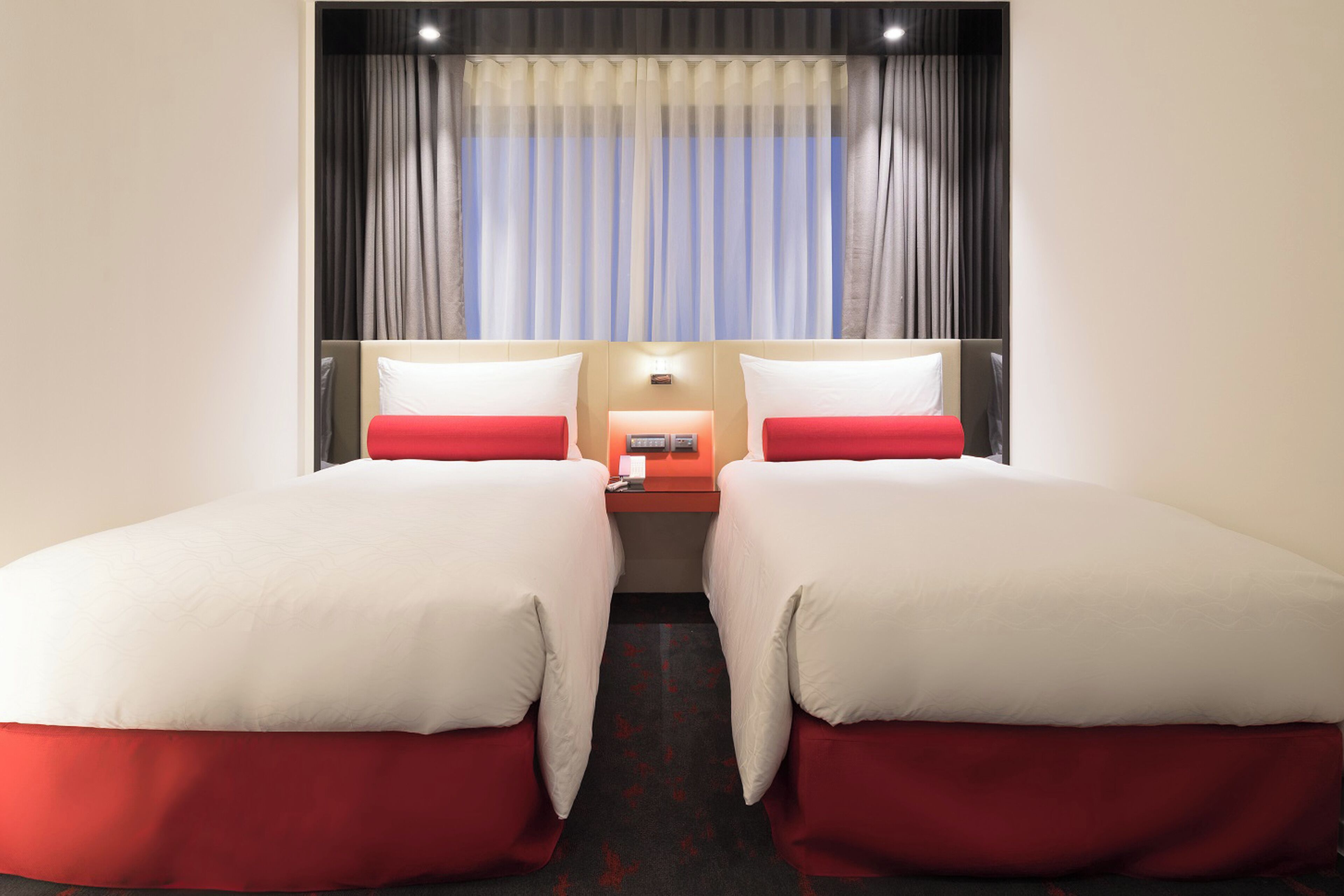 A hotel room with two beds, a nightstand, a lamp, and a window with curtains.