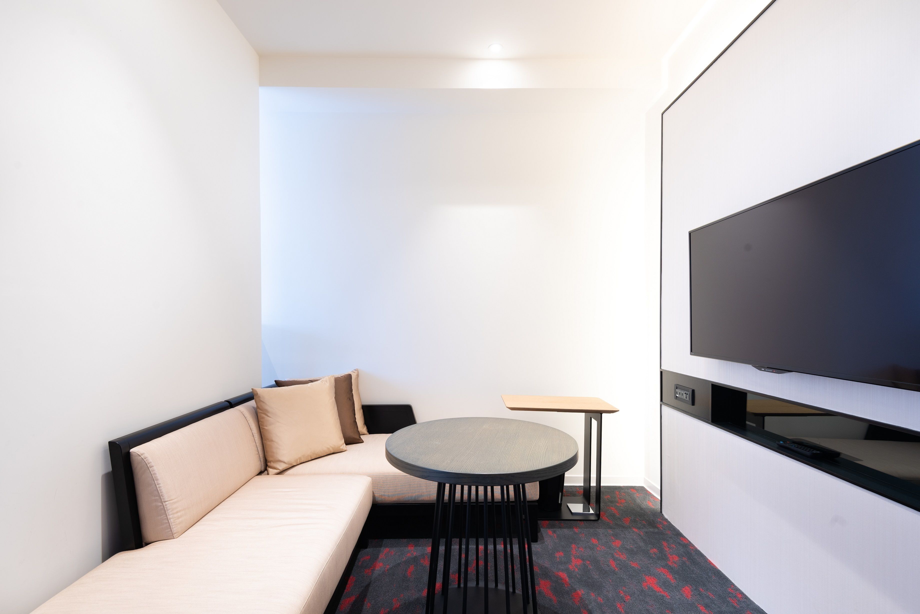 A compact hotel room with a sofa, a small round table, and a flat-screen TV mounted on the wall.