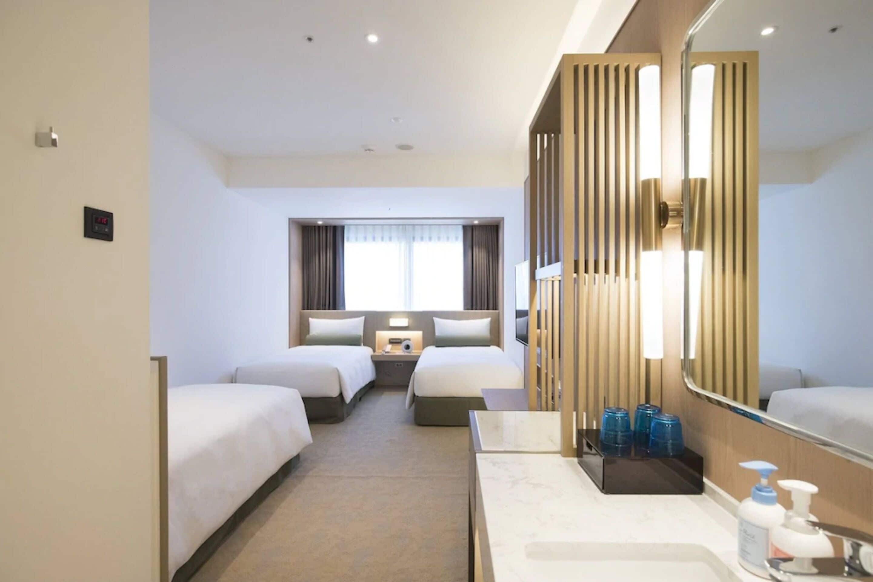 Habitación de hotel con dos camas, un espejo grande, baño con lavamanos y artículos de aseo, y vista a la calle.