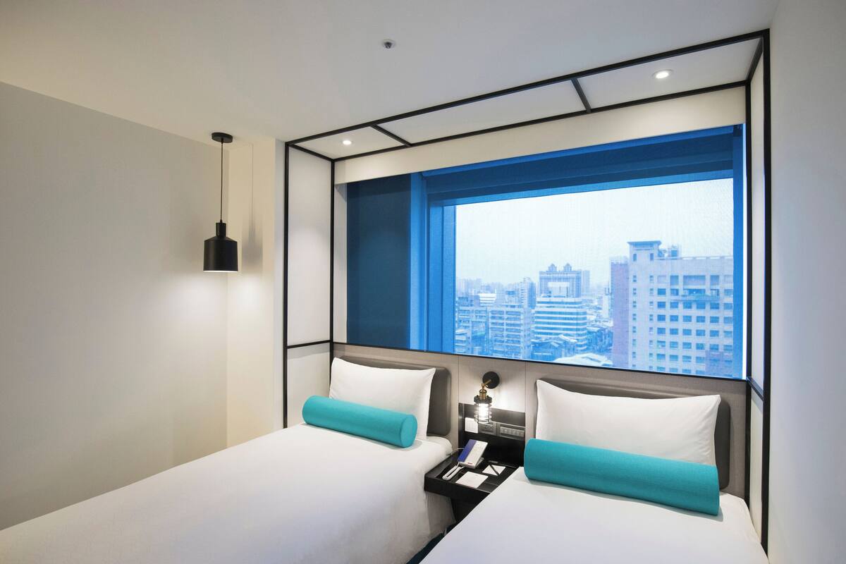 A modern hotel room with a large window, a bed, a bedside table, and a hanging light fixture.
