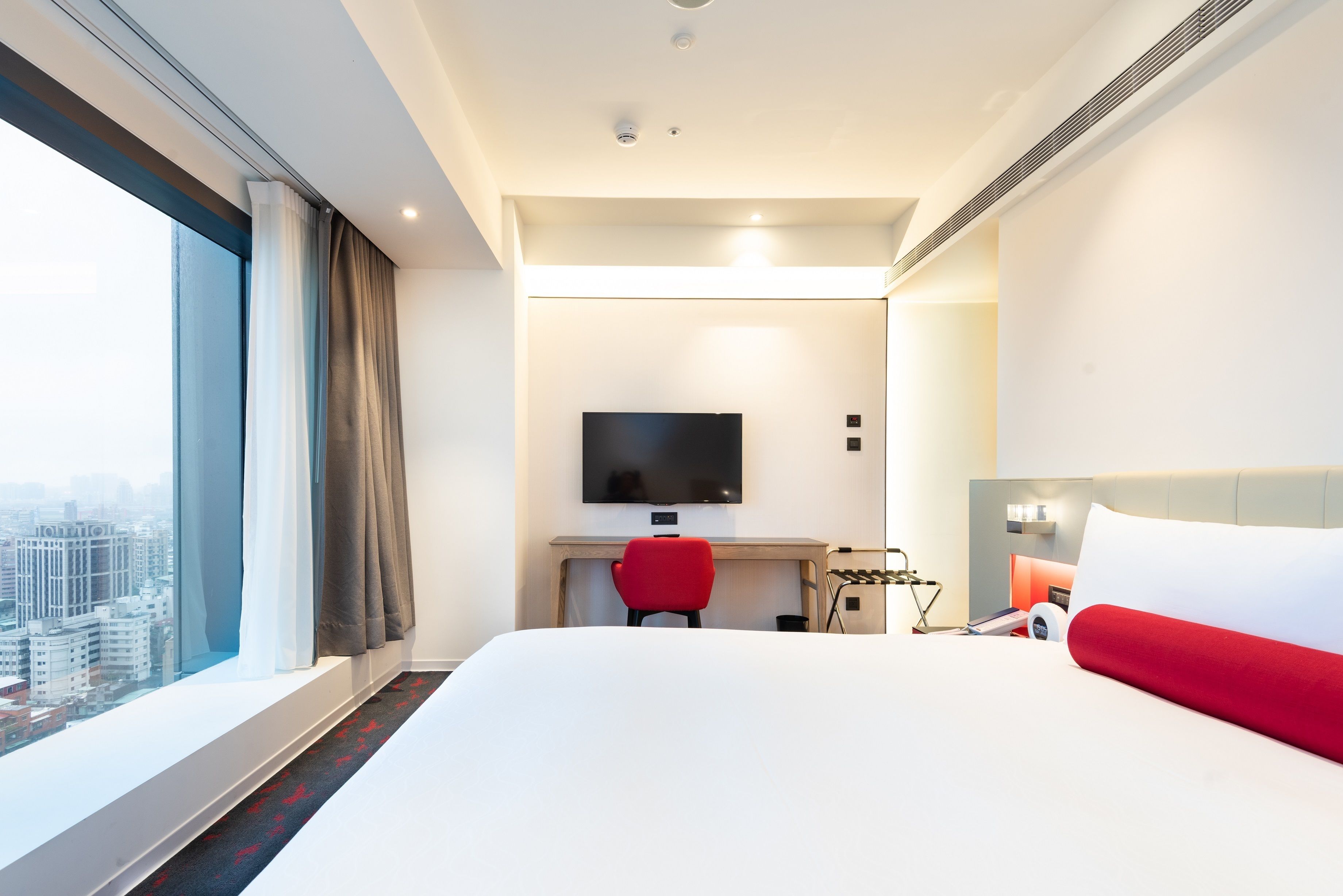 A hotel room with a large bed, a desk with a chair, a TV, and a city view.