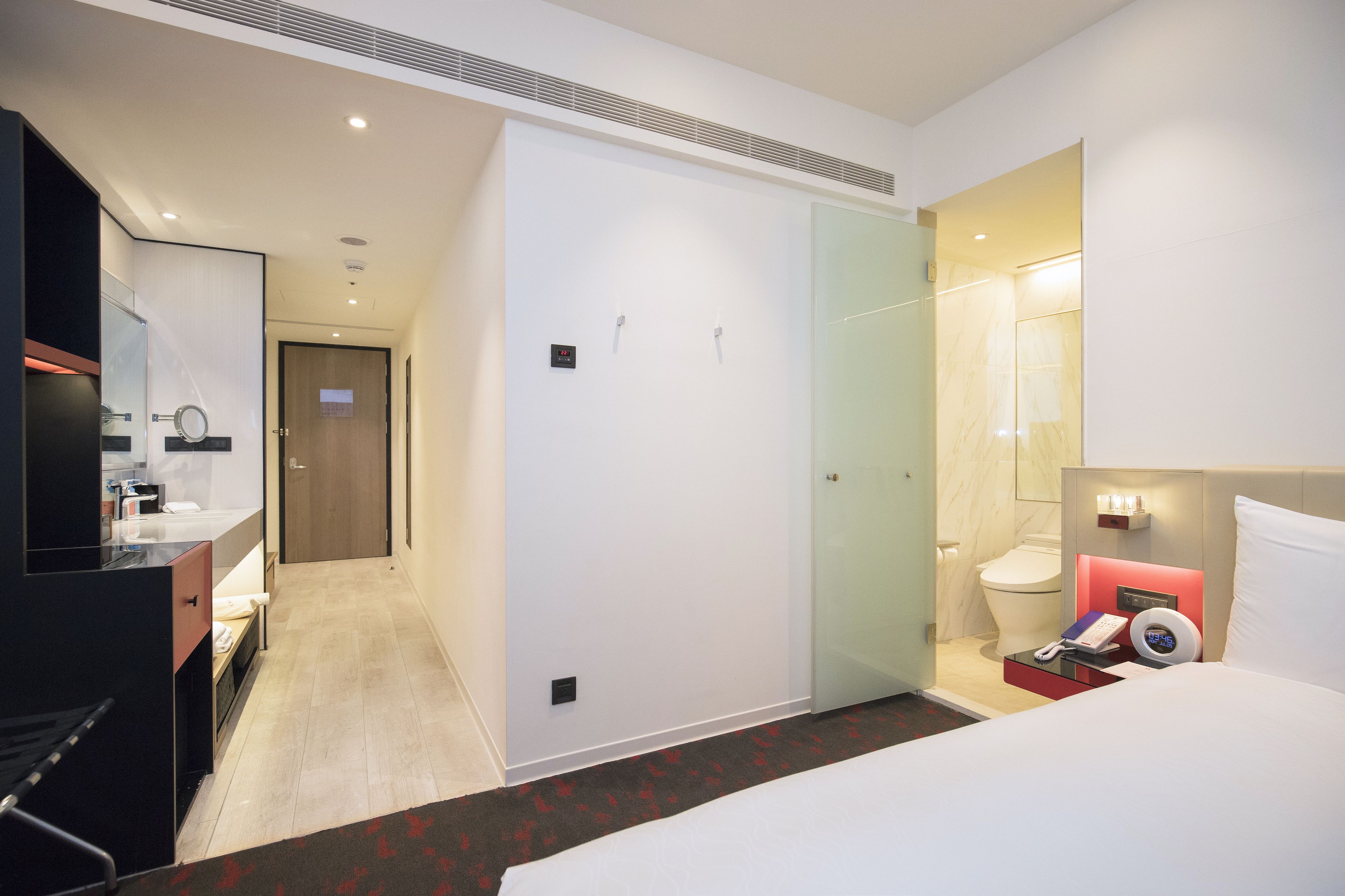 A hotel room with a bed, a bedside table with a phone and a remote, a mirror, a wardrobe, a bathroom with a toilet and a shower, and a hallway leading to another room.