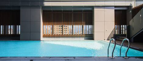 Una piscina con cubierta de madera, barandales de metal y vistas a edificios al fondo.
