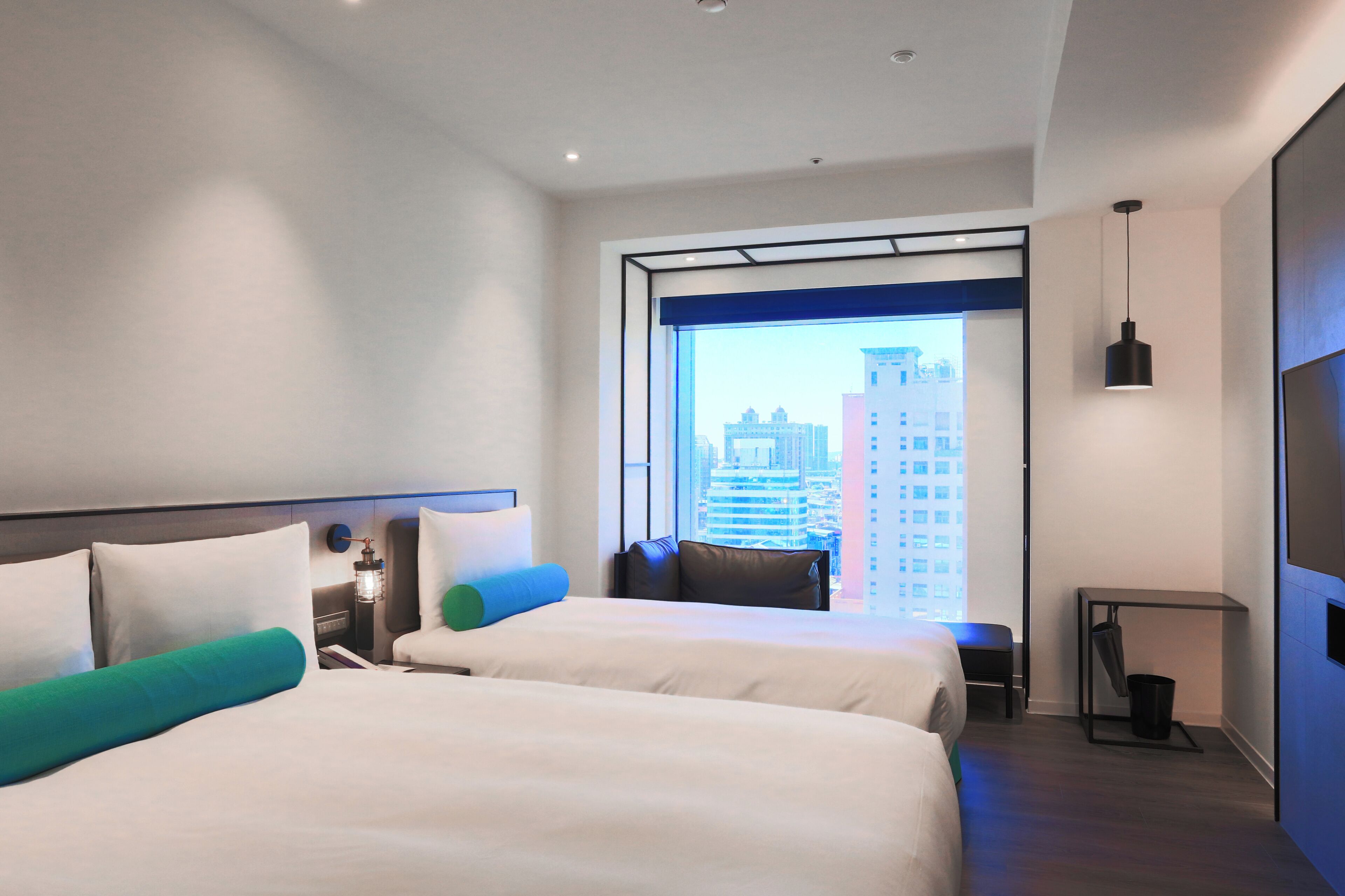 Quarto de hotel moderno com duas camas, vista para a cidade, uma mesinha e um lustre.