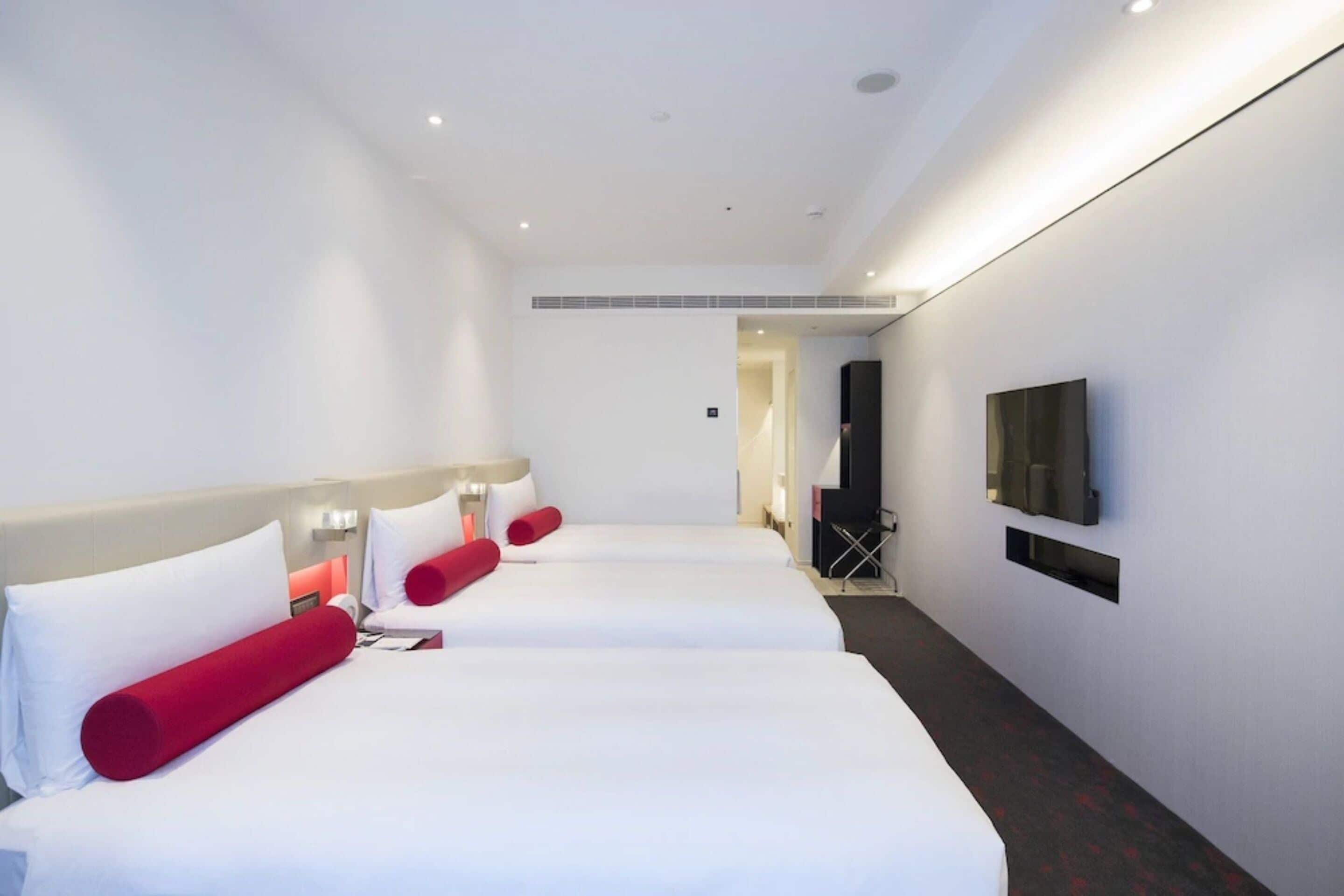 Habitación de hotel con tres camas, un televisor montado en la pared y un pasillo que conduce a otra habitación.