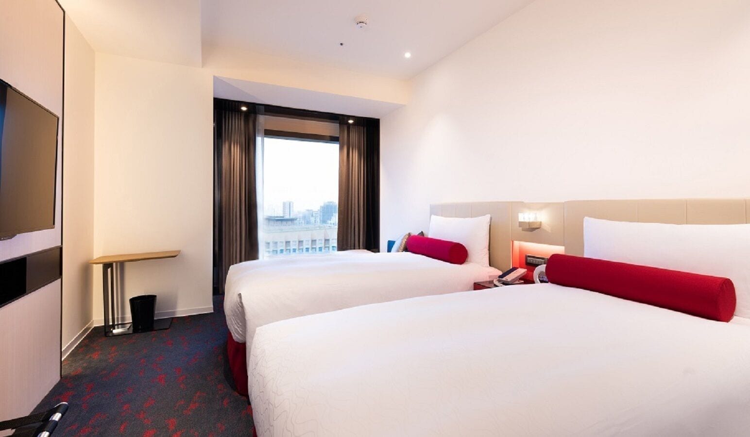 A hotel room with two beds, a TV, a desk, and a window with a city view.