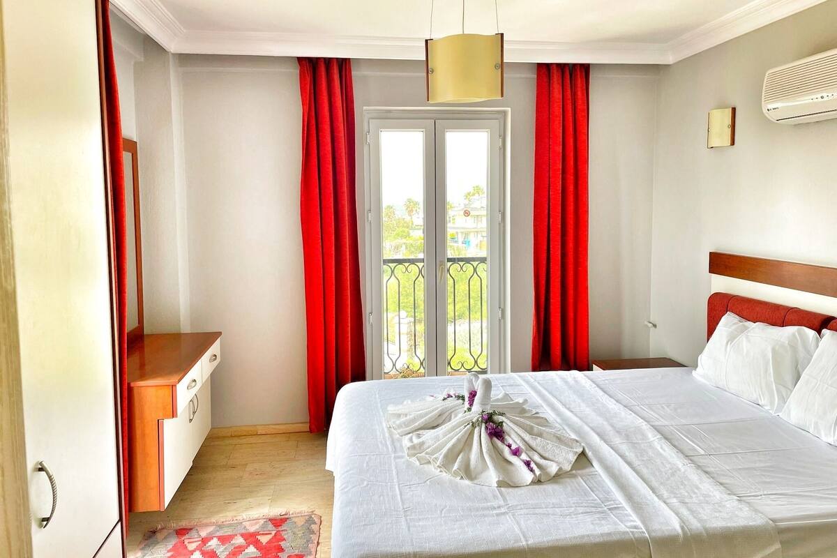 A hotel room with a bed, a desk, a chair, a window with red curtains, and a balcony.