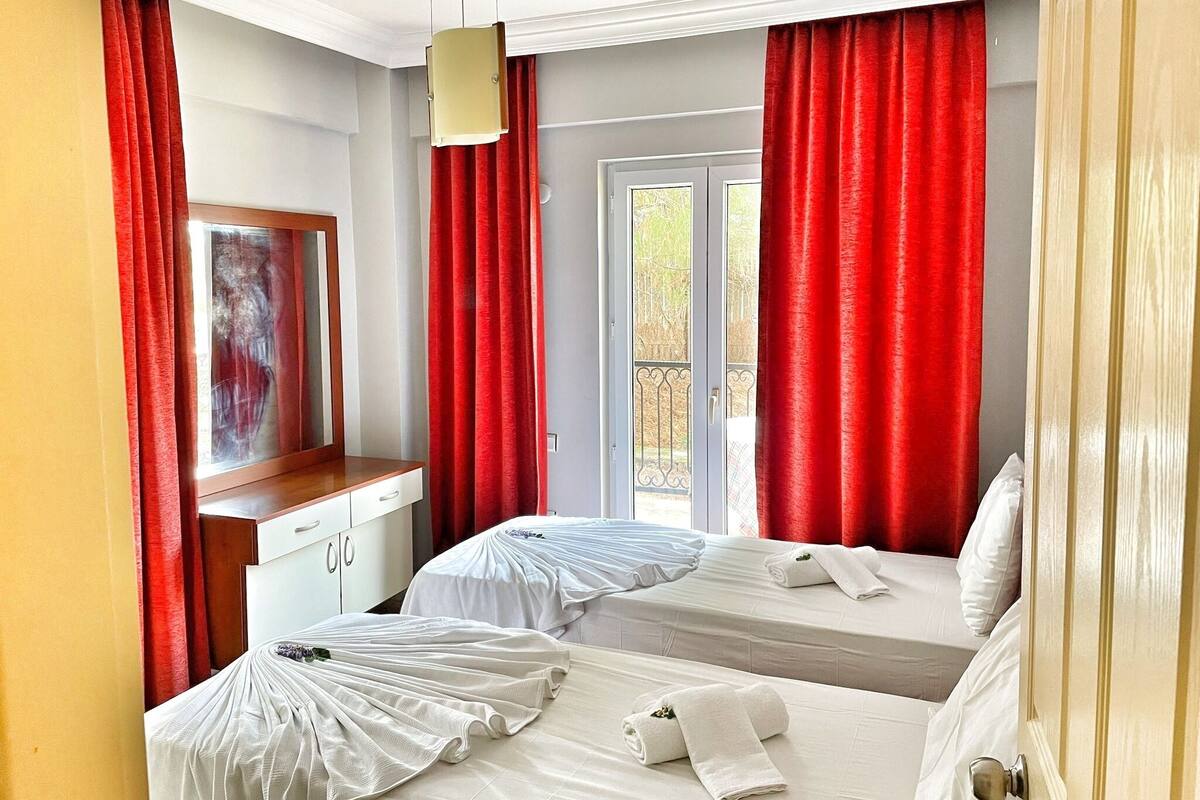 A hotel room with two beds, red curtains, a small desk with a mirror, and a window with a view of trees.