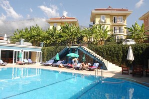 A swimming pool with lounge chairs and umbrellas, surrounded by buildings and greenery. - Antas Deluxe Apartments (Fethiye)