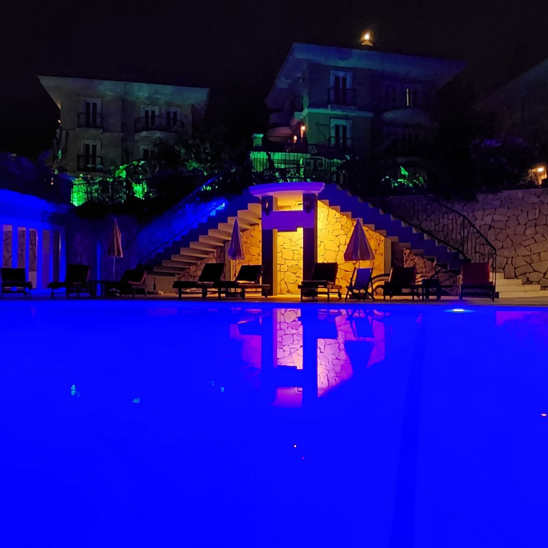 A nighttime swimming pool area with a stone wall and a staircase leading to a building.