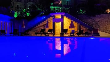 A nighttime swimming pool area with a stone wall and a staircase leading to a building.