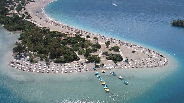 An aerial view of a beach resort with rows of sunbeds, a small island with boats, and a clear blue sea. - Antas Deluxe Apartments (Fethiye)