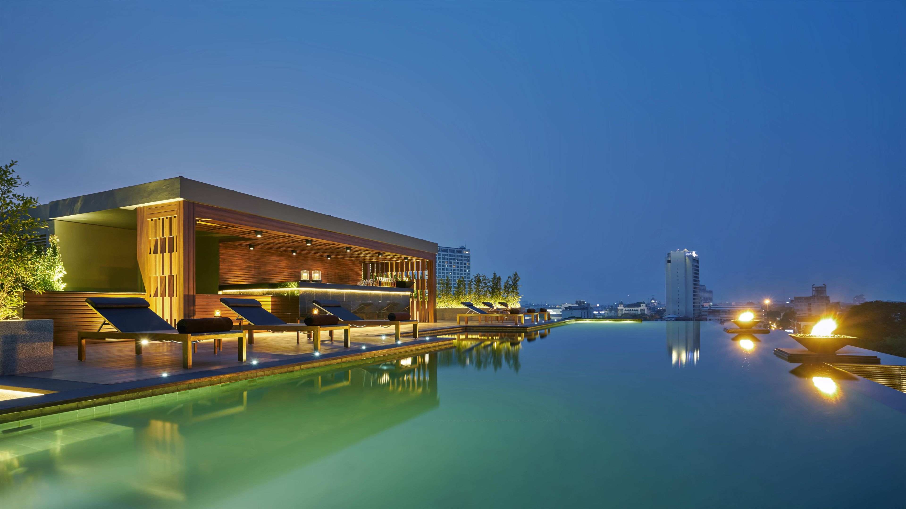 A modern poolside area with lounge chairs and a bar, surrounded by a calm pool and city skyline.