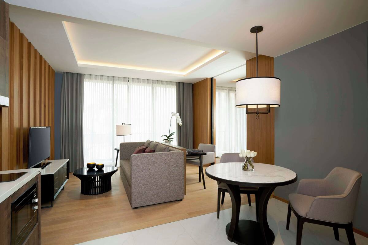 Une chambre d’hôtel moderne dotée d’une télévision à écran plat, d’un canapé, d’une table à manger avec des chaises et d’une grande fenêtre avec des rideaux.