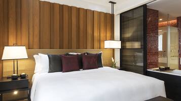 A hotel room with a large bed, two bedside tables with lamps, a wooden panel wall, and a glass shower enclosure.