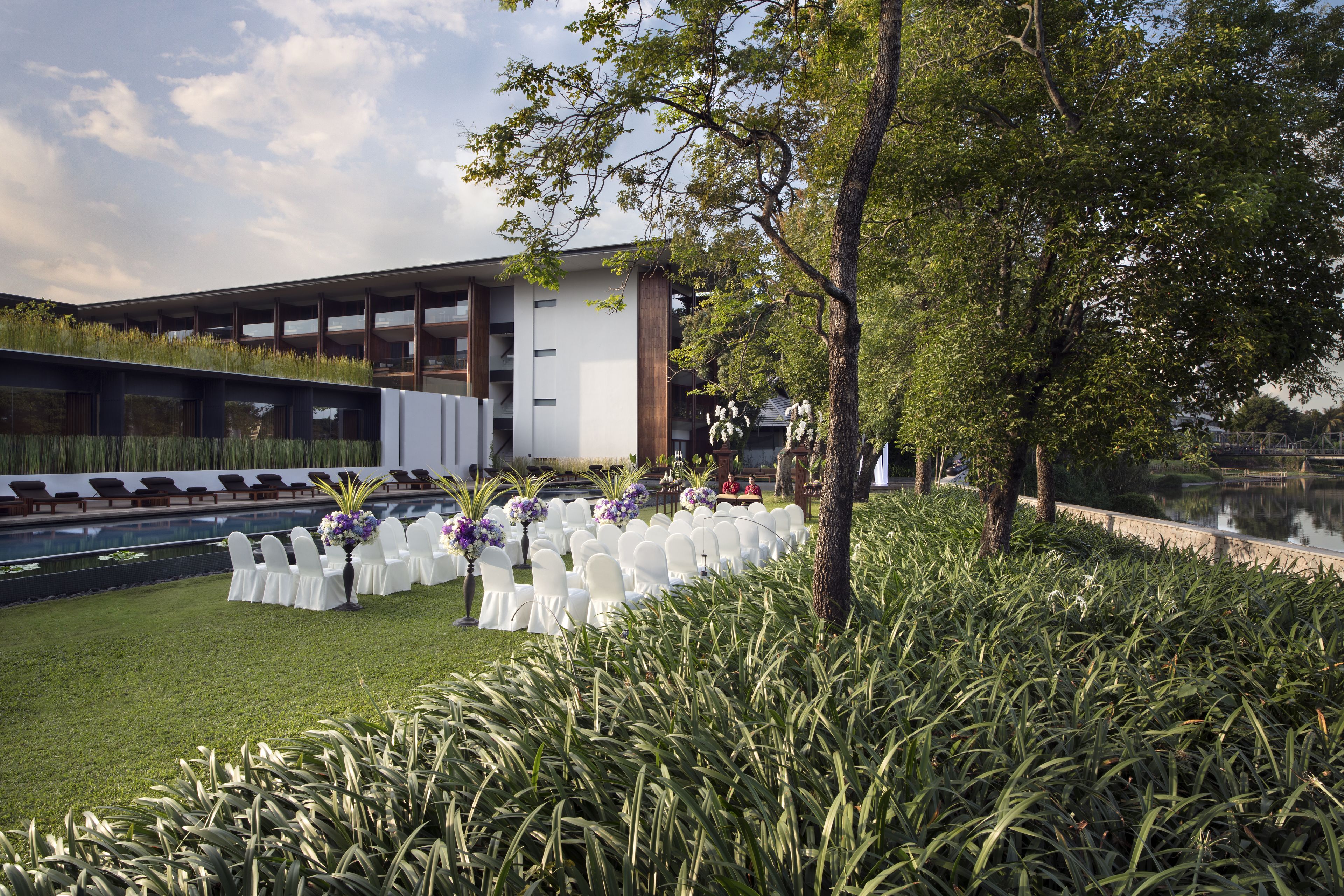 A modern building with a pool, surrounded by lush greenery and white chairs arranged for an event.