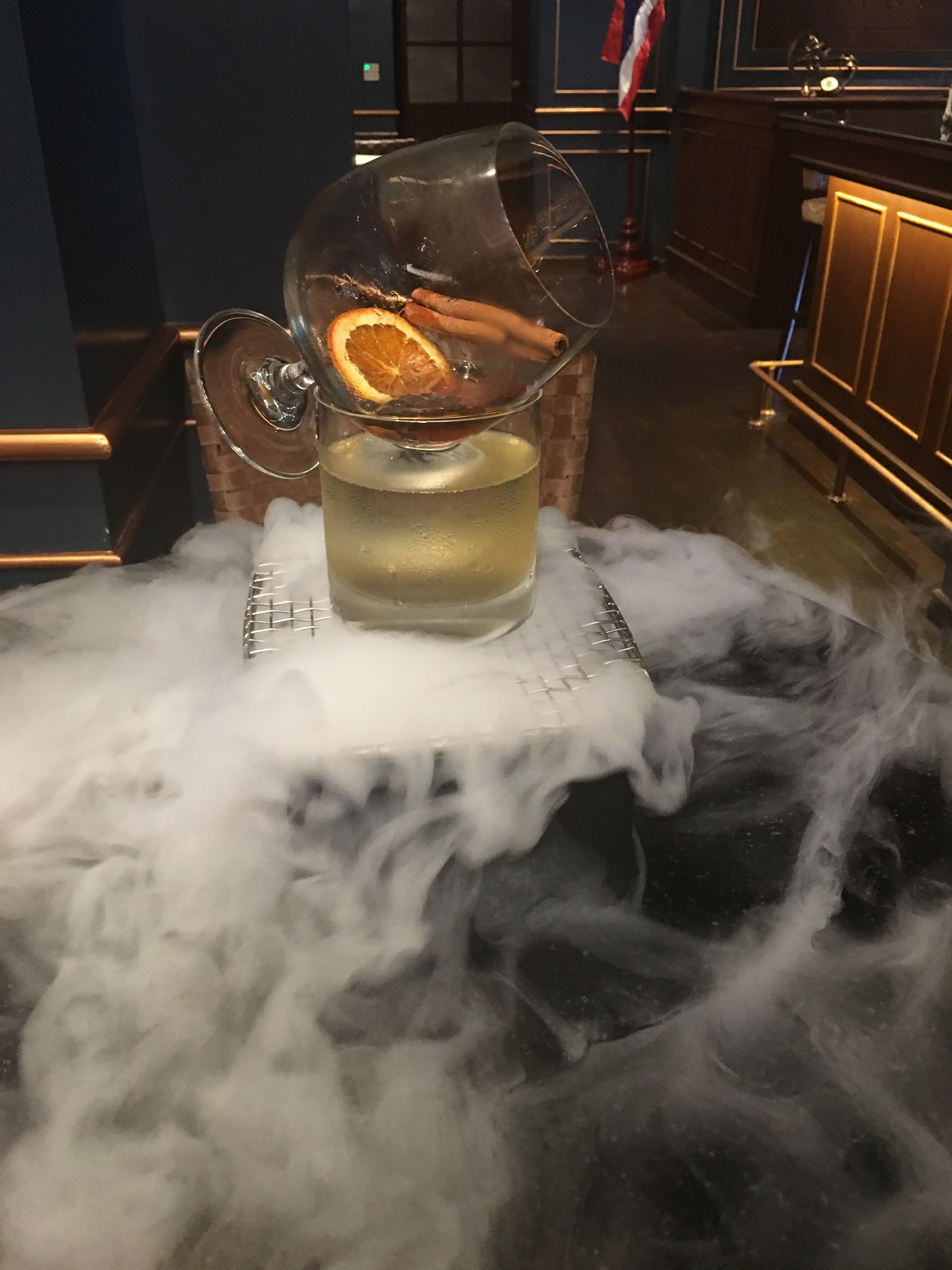 Un vaso de líquido con tapa transparente tipo cúpula, una rodaja de naranja y palitos de canela sobre una barra con un mantel blanco.
