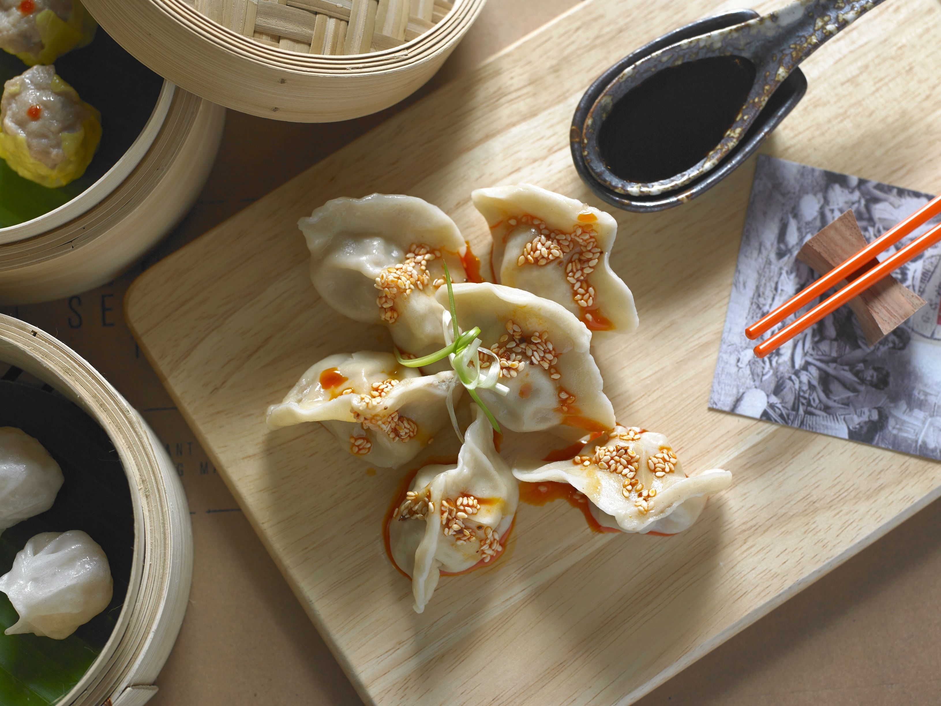 A wooden table with a plate of dumplings garnished with sesame seeds and green onions, accompanied by a small bowl of soy sauce and chopsticks.