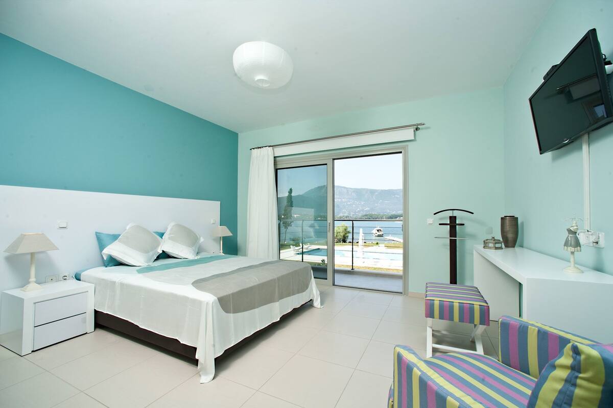Un dormitorio con cama, mesitas de noche, televisor, sillón y vistas a una piscina y montañas.