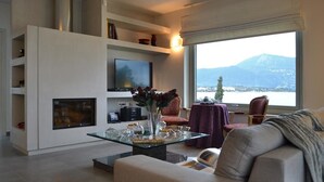 A modern living room with a glass coffee table, a sofa, a TV mounted on the wall, and a view of a mountain and lake through large windows. - K&K Luxury Beach Villas (Corfu)