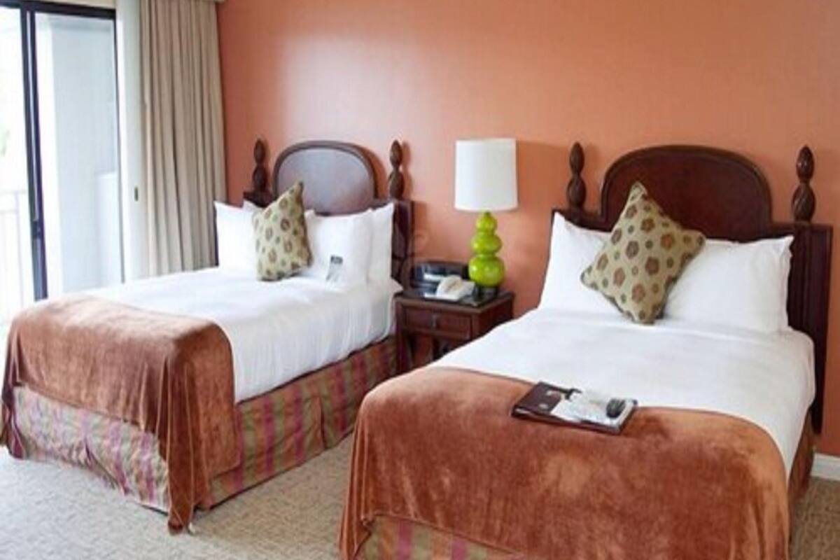A hotel room with two beds, each with a headboard, a bedside table with a lamp, and a window with curtains.