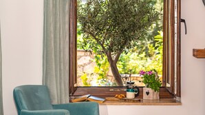 A room with a window overlooking a garden, a teal armchair, a wooden shelf with a coffee maker, and a book.