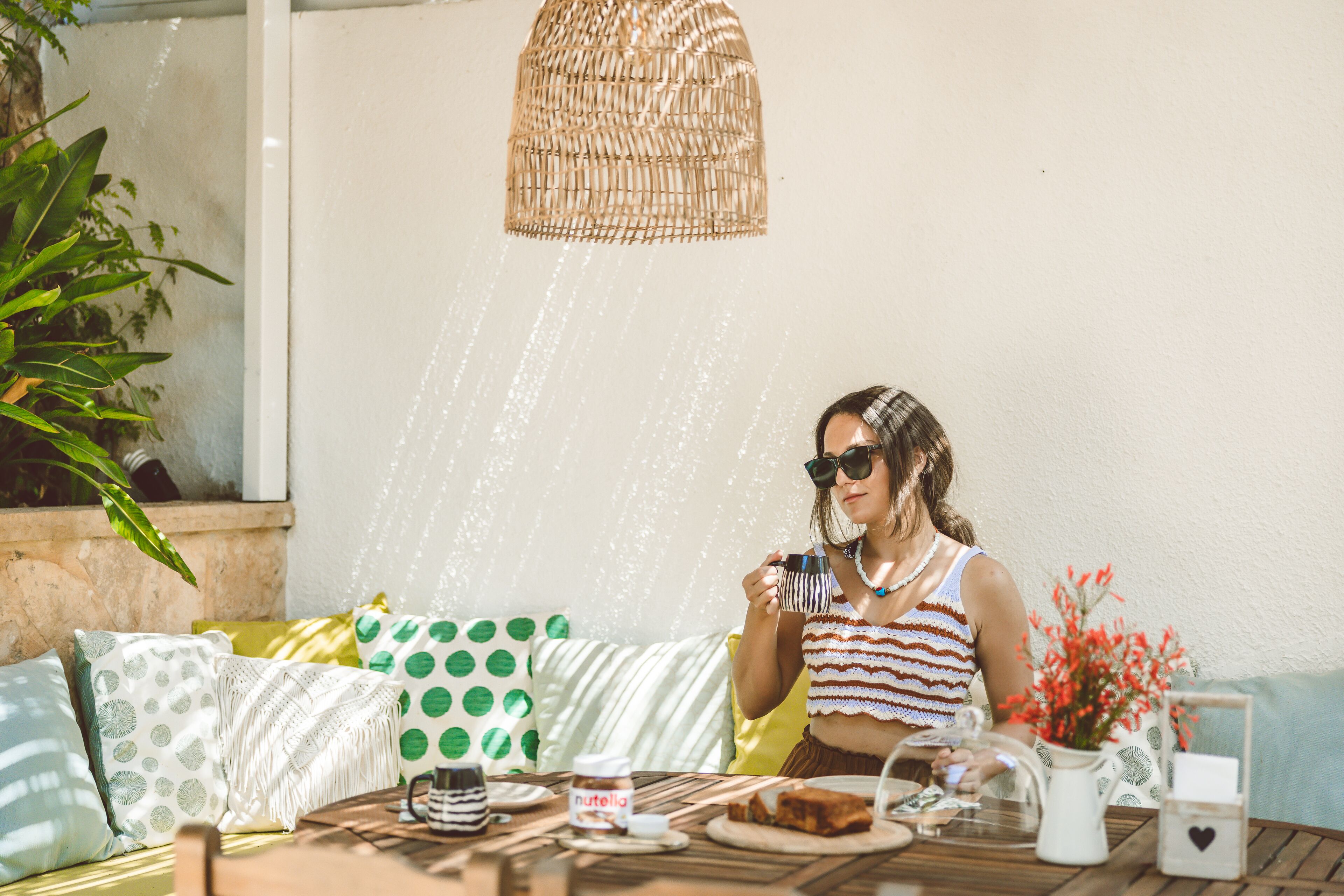 A person sitting at an outdoor table with a wicker lamp, various cushions, a jar of Nutella, and a glass of water.