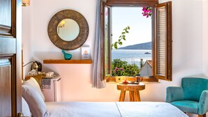 A bedroom with a bed, a window with a view of the sea, a chair, and a small table with a lamp.