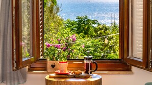 A window with a view of the sea and greenery, a coffee pot, a cup, and a plate with cookies on a wooden table.