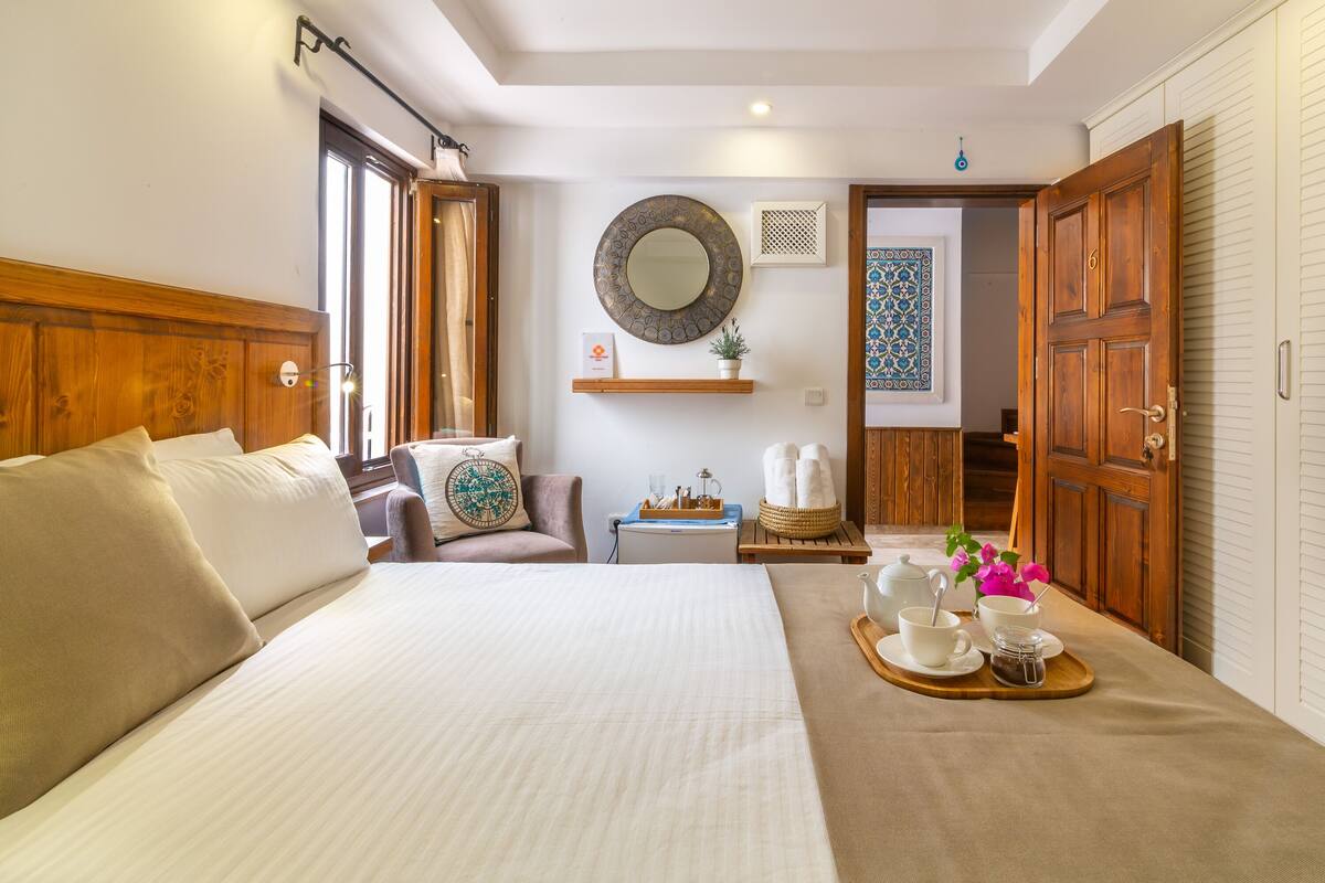 Une chambre d’hôtel avec un grand lit, une table de chevet sur laquelle se trouve une théière et des tasses, un miroir et une armoire en bois.