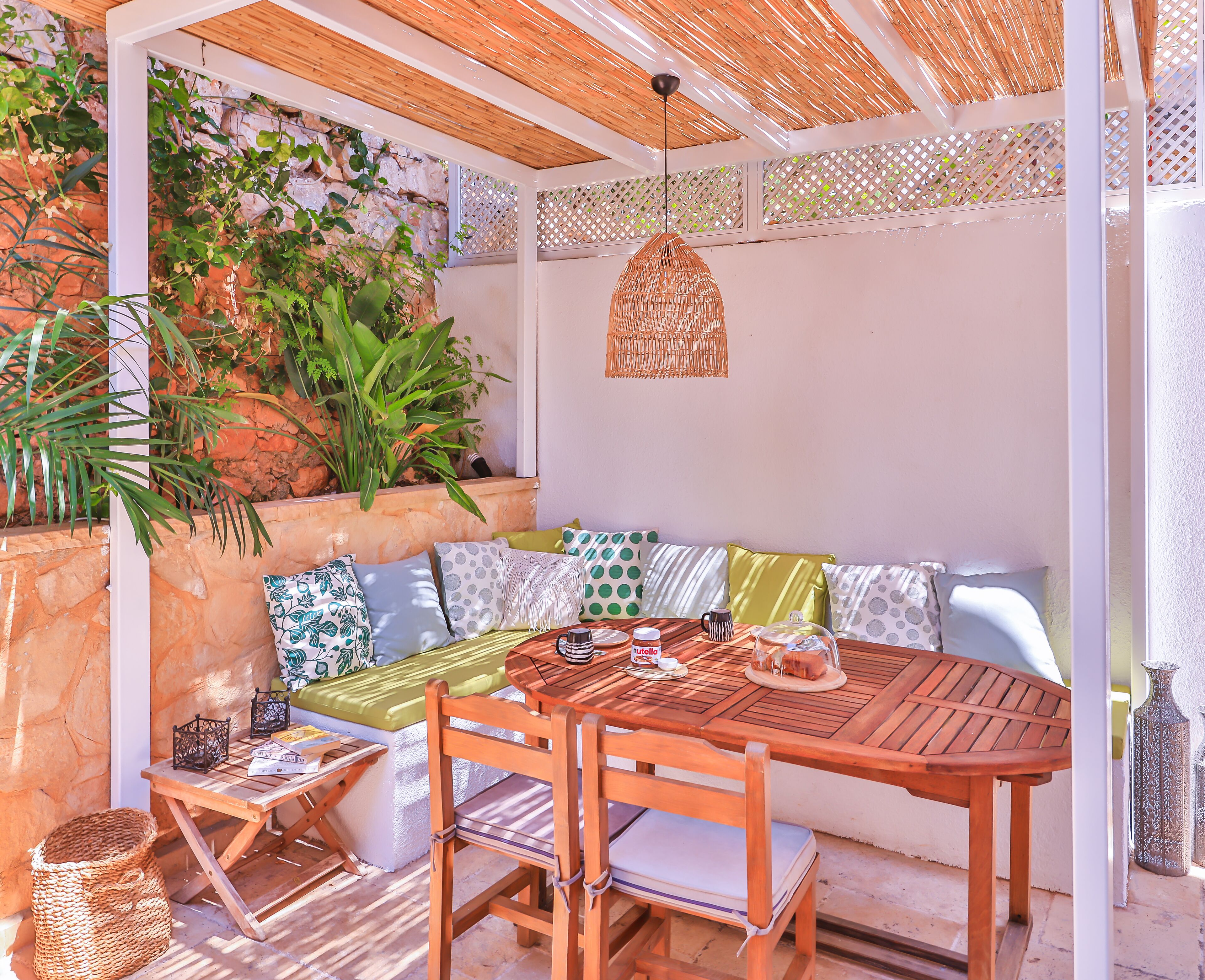 Une terrasse couverte avec une table en bois, des chaises et un banc agrémenté de coussins. On y trouve également des plantes et un luminaire suspendu.